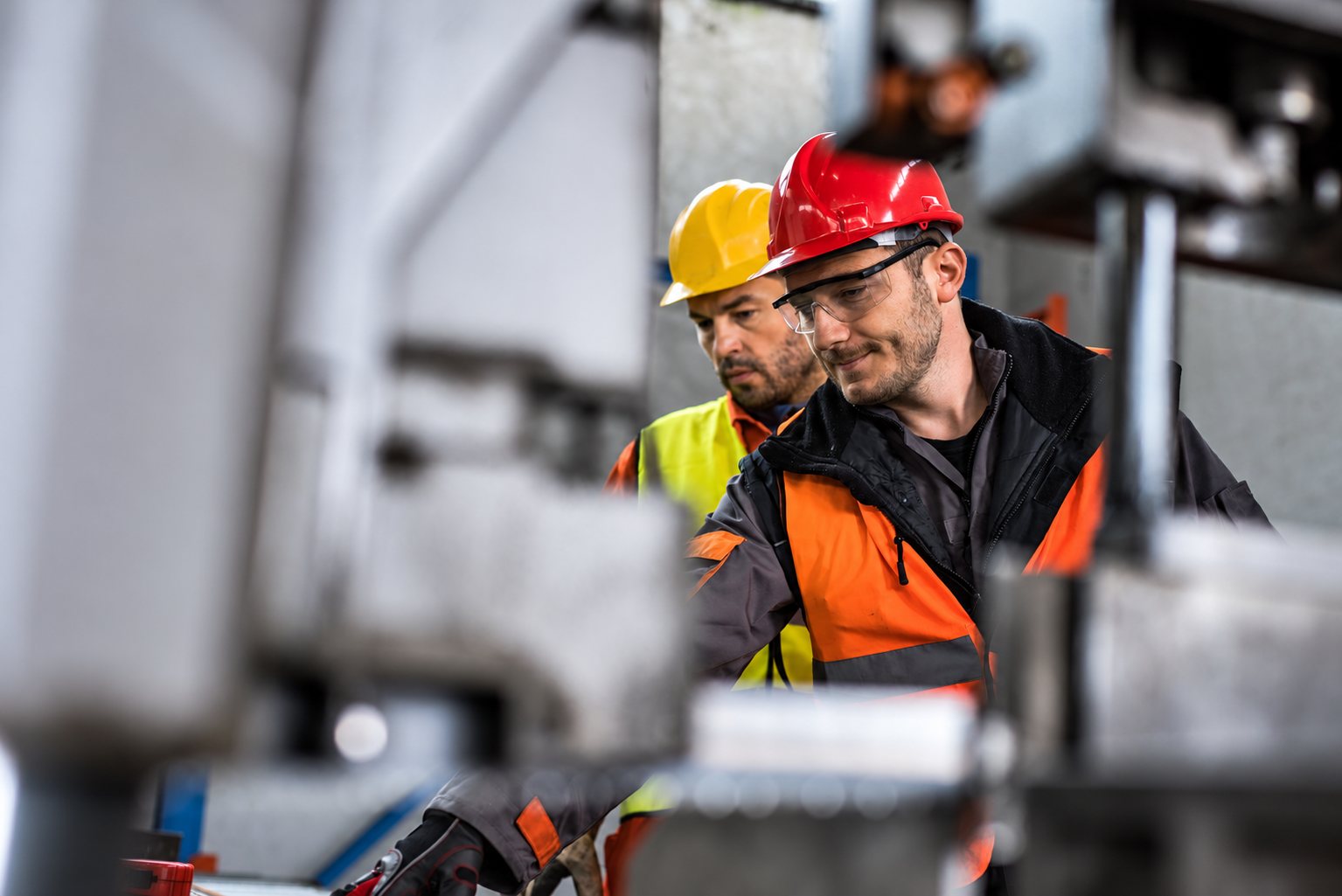 Deux ouvriers portant casque, lunettes de protection et gilets de sécurité orange et jaune travaillent près de machines industrielles.