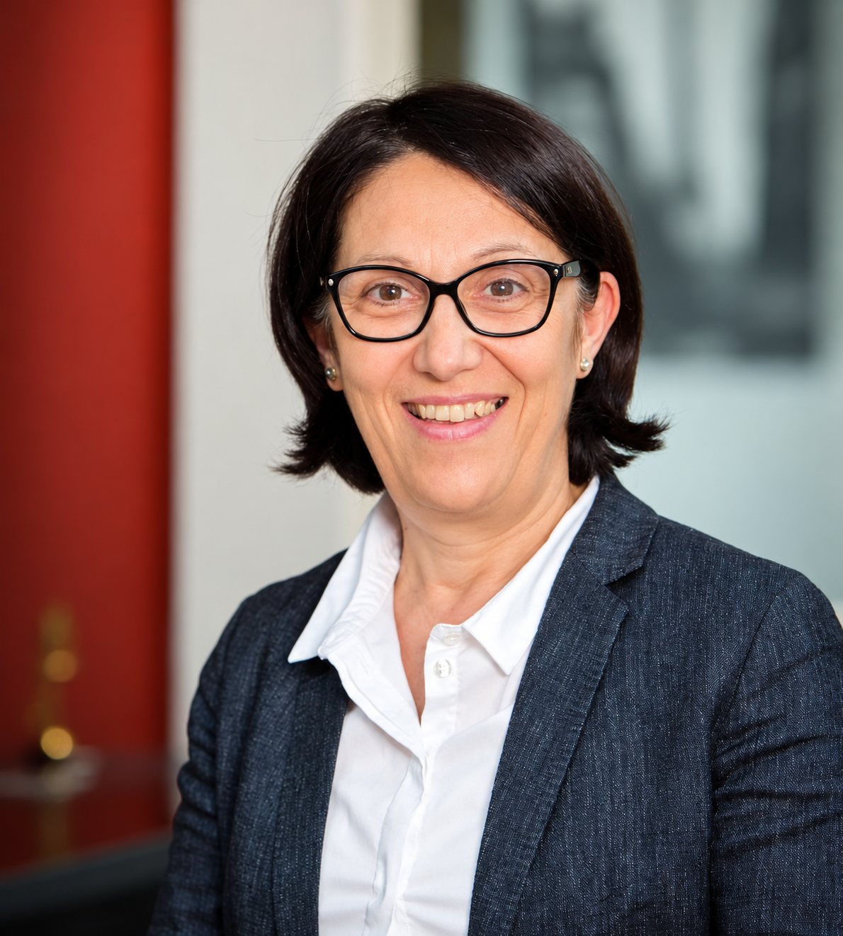Portrait d’Eladia Delgado, souriante, cheveux bruns courts, lunettes noires et veste grise.