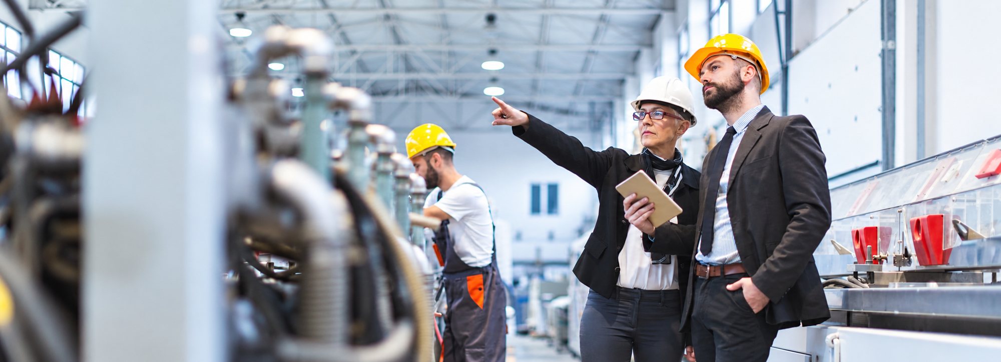 Dans une grande usine, une femme et deux hommes portant des casques observent une chaîne de production.